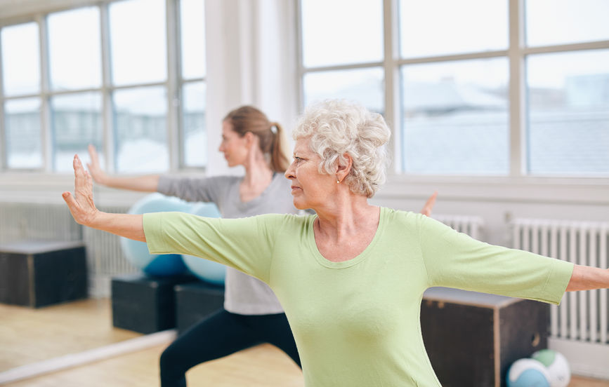 two women doing stretching and yoga workout at gym. female trainer in background with senior woman in front during physical training session