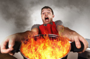 45948776 - young inexperienced home cook with apron holding pot burning in flames with stress and panic face expression in fire in the kitchen and cooking wrong concept