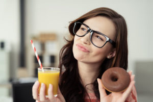 39184009 - charismatic young woman wearing glasses deciding between a healthy and unhealthy diet as she holds up a chocolate doughnut an glass of fresh orange juice