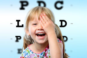 47634708 - close up face portrait of happy girl having fun at vision test.conceptual image with girl closing one eye with hand and block letter eye chart in background.