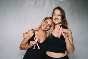 Two women, friends, hugging each other and holding up pink ribbons to show solidarity for breast cancer awareness.