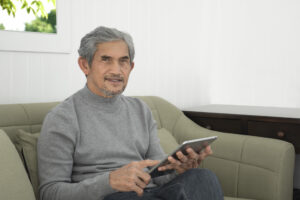 Portrait elderly senior asian man sits near glass window in the morning to work from home and checking his business on his laptop on table seriously, soft and selective focus