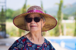 Portrait of senior woman wearing sunglasses and straw hat while standing side swimming pool. Space for text. Concept of aged people and relaxation.