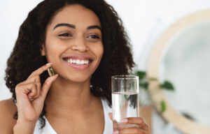 Daily Intake Of Vitamins And Minerals. Happy Black Woman Holding Capsule With Beauty Supplements And Water Glass, Closeup