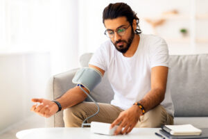 Young man sitting on couch, checking blood pressure and taking notes at home. Indian guy in white t-shirt using modern tonometer, experiencing hypertension caused by stress, copy space