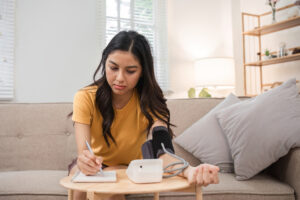 Young woman checks her health at home using a blood pressure monitor, taking notes in a cozy living room environment.