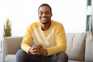 Happy african american guy sitting on couch in grey living room, smiling at camera, copy space. Closeup of positive young black man chilling on sofa, weekend at home concept
