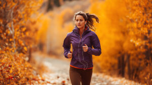 A focused woman in a purple jacket jogs on a trail, surrounded by the vibrant golden-orange foliage of an autumn forest while listening to music on her earphones.