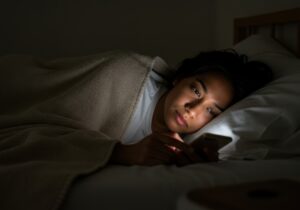 A young woman lies in bed at night, engrossed in her smartphone, the screen casting a soft light on her face as she scrolls through content.