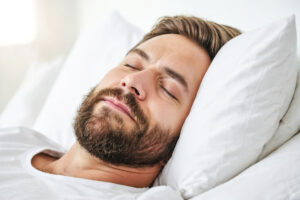 A young man sleeping soundly in a bed with white bedding. He is resting peacefully, suggesting a good night's sleep. This image can be used for stock photos or for a blog about sleep or relaxation.